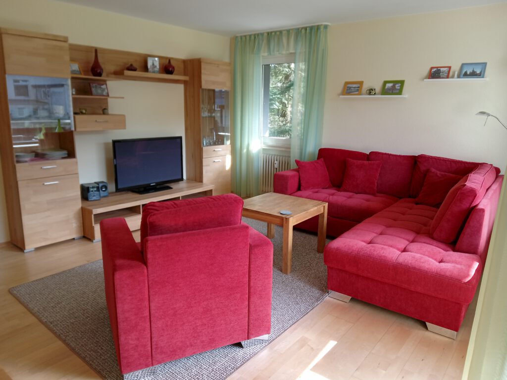Das helle Wohnzimmer ist in warmen Farbtönen gestaltet. Ein rotes Sofa und ein passender Sessel stehen vor einem Flachbild-TV. Ein Holztisch dient als Mittelpunkt, während ein gemusteter Teppich den Boden schmückt.